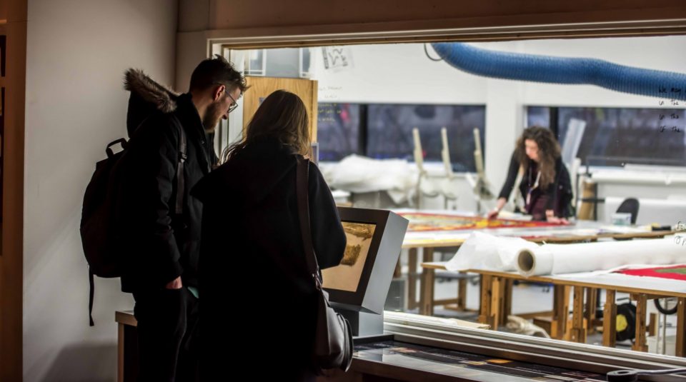 Image of Visitors in front of the viewing window into The Conservation Studio, where a conservator is on a table working on a banner.