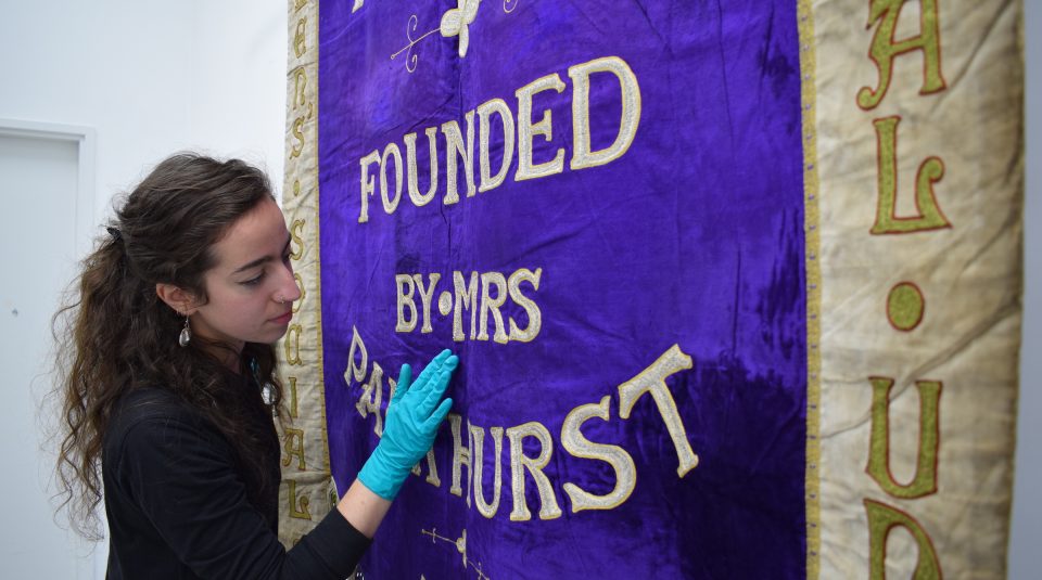 Image of Senior Conservator Kloe Rumsey with the 1908 Manchester suffragette banner in The Conservation Studio at People’s History Museum.