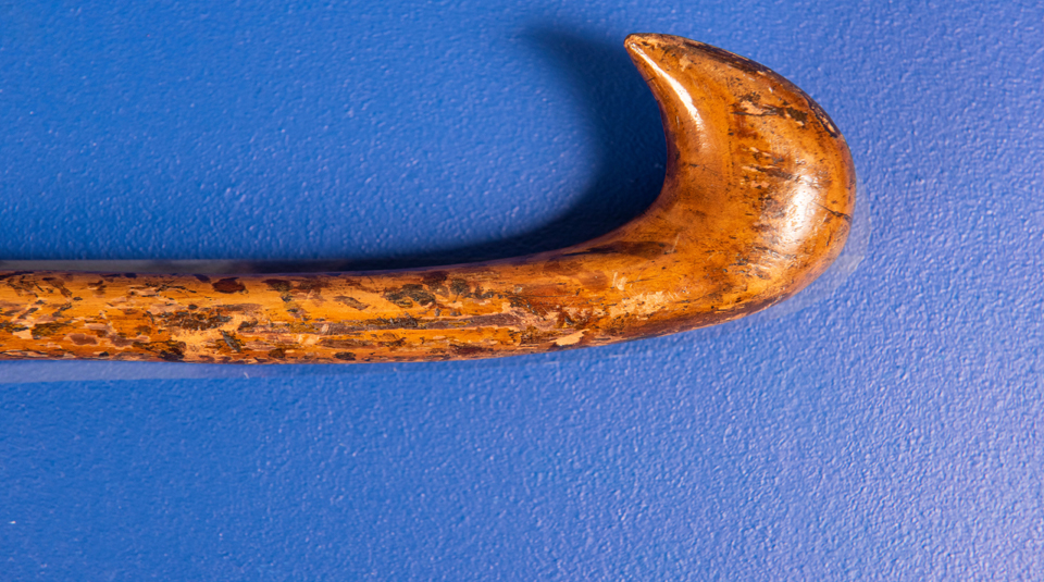 Image of The top of a brown wooden cane on a blue background, showing an engraving on the cane of a cap of liberty.