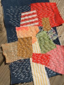 Image shows a patchwork textile piece, it has been made from different colours of material in red, pink, blue, green, brown and white. There is white stitching over the patchwork in vertical lines. 