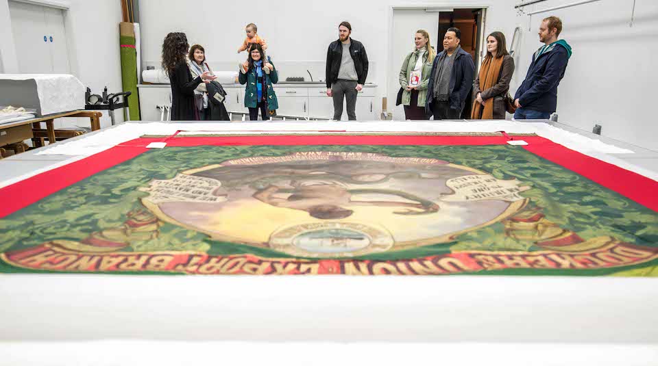 Image of A group of people listening to a member of museum staff, standing near a huge historic banner laid out on a table.
