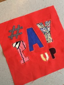 Red textile square with text #PAY UP. The Fabric of Protest workshop, 10 December 2022, image courtesy of Helen Mather for People's History Museum