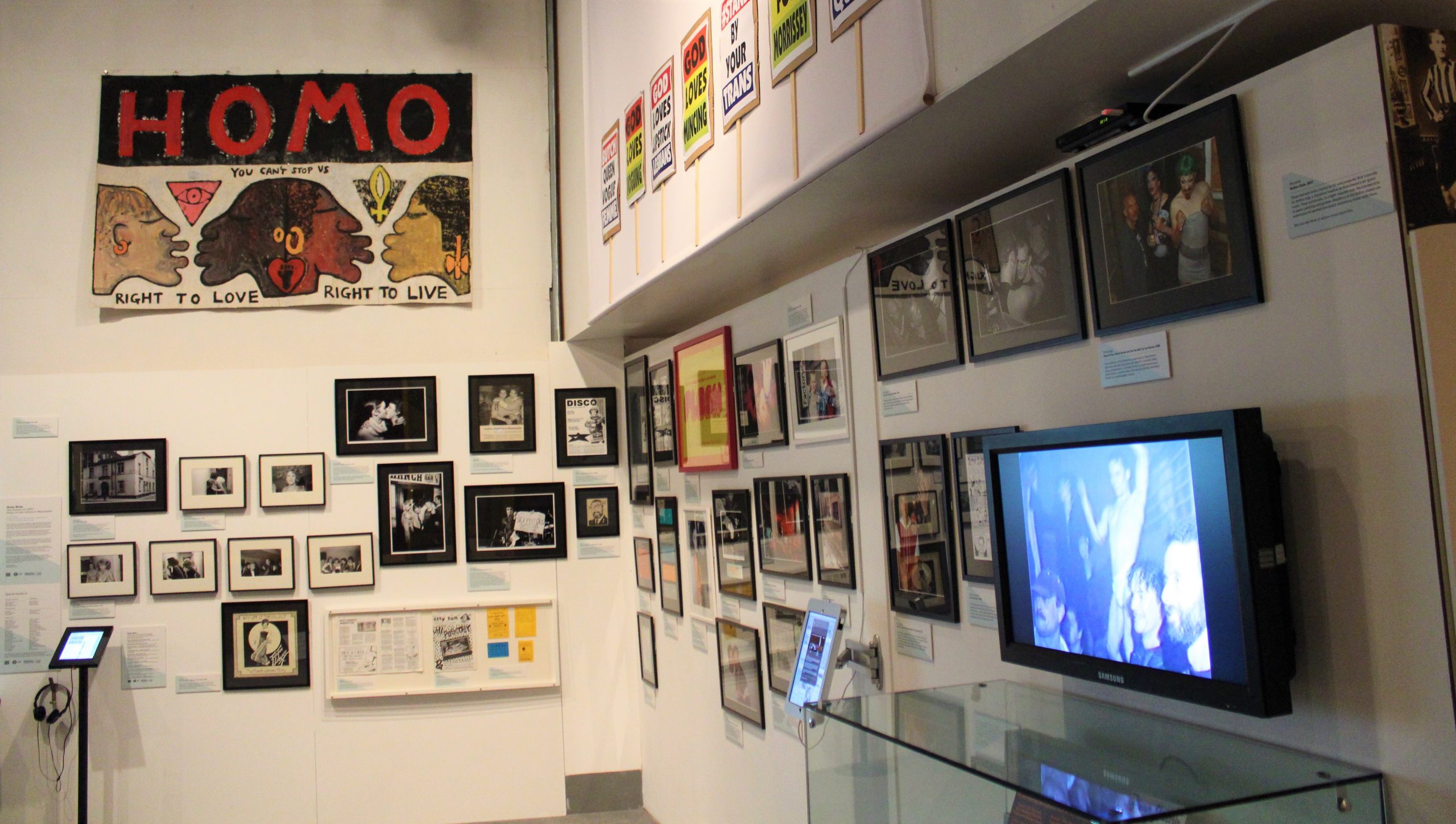 Queer Noise exhibition at People's History Museum, 2017. Photograph of the corner of an L shaped exhibition wall covered with framed photographs, images and a television monitor. At the top of the far wall is a large painted paper banner which includes the profiles of four kissing faces, each is a different colour. The banner reads ‘HOMO - You Can’t Stop Us - Right To Love - Right To Live’.