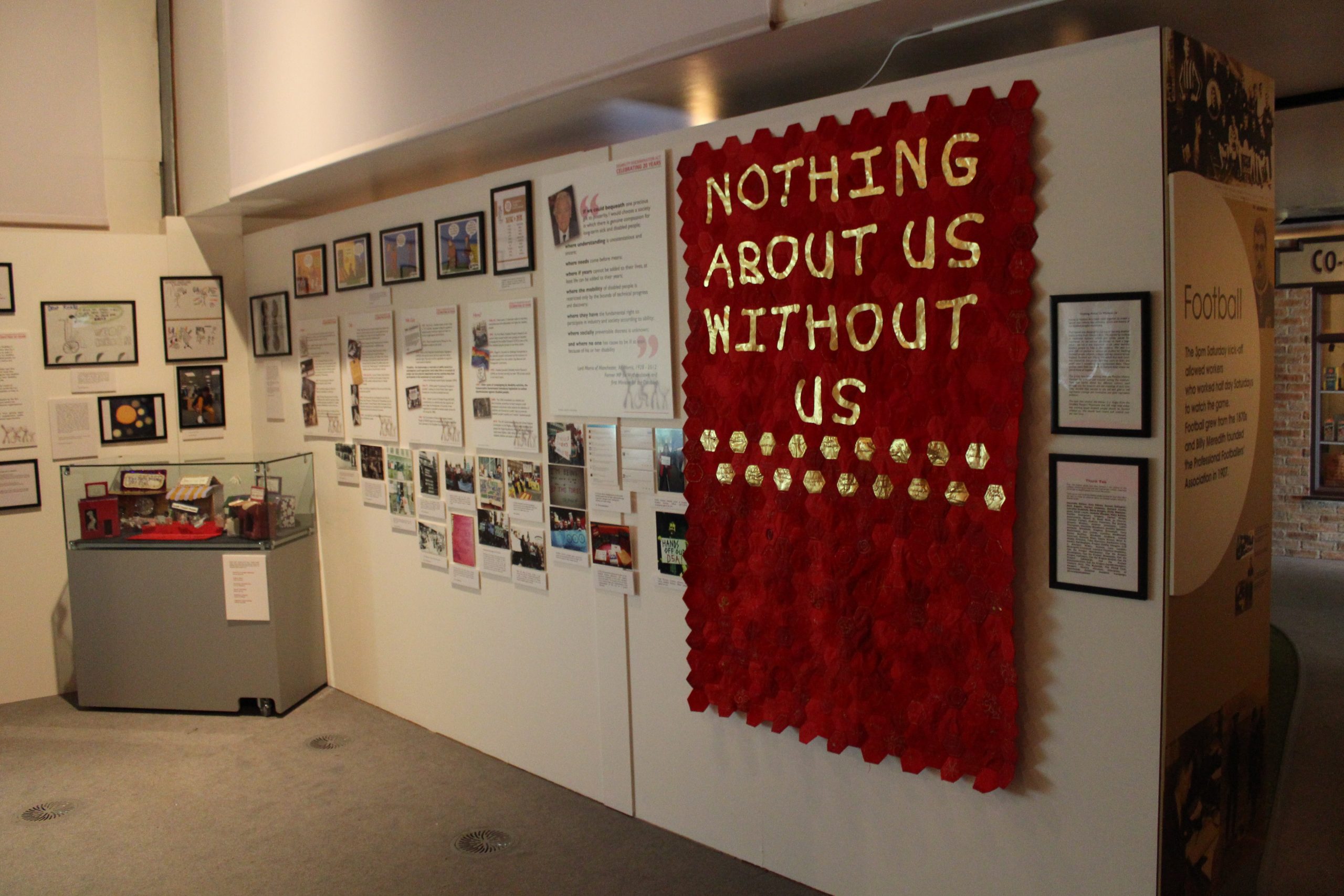 The Disability Discrimination Act, Celebrating 20 Years exhibition at People's History Museum, 2015. Photograph of an L shaped exhibition wall covered with images and text panels. At the right end of the wall is a red banner made up of small hexagons of material with gold lettering that reads ‘Nothing About Us Without Us’. At the left end of the wall is a glass case containing small objects.