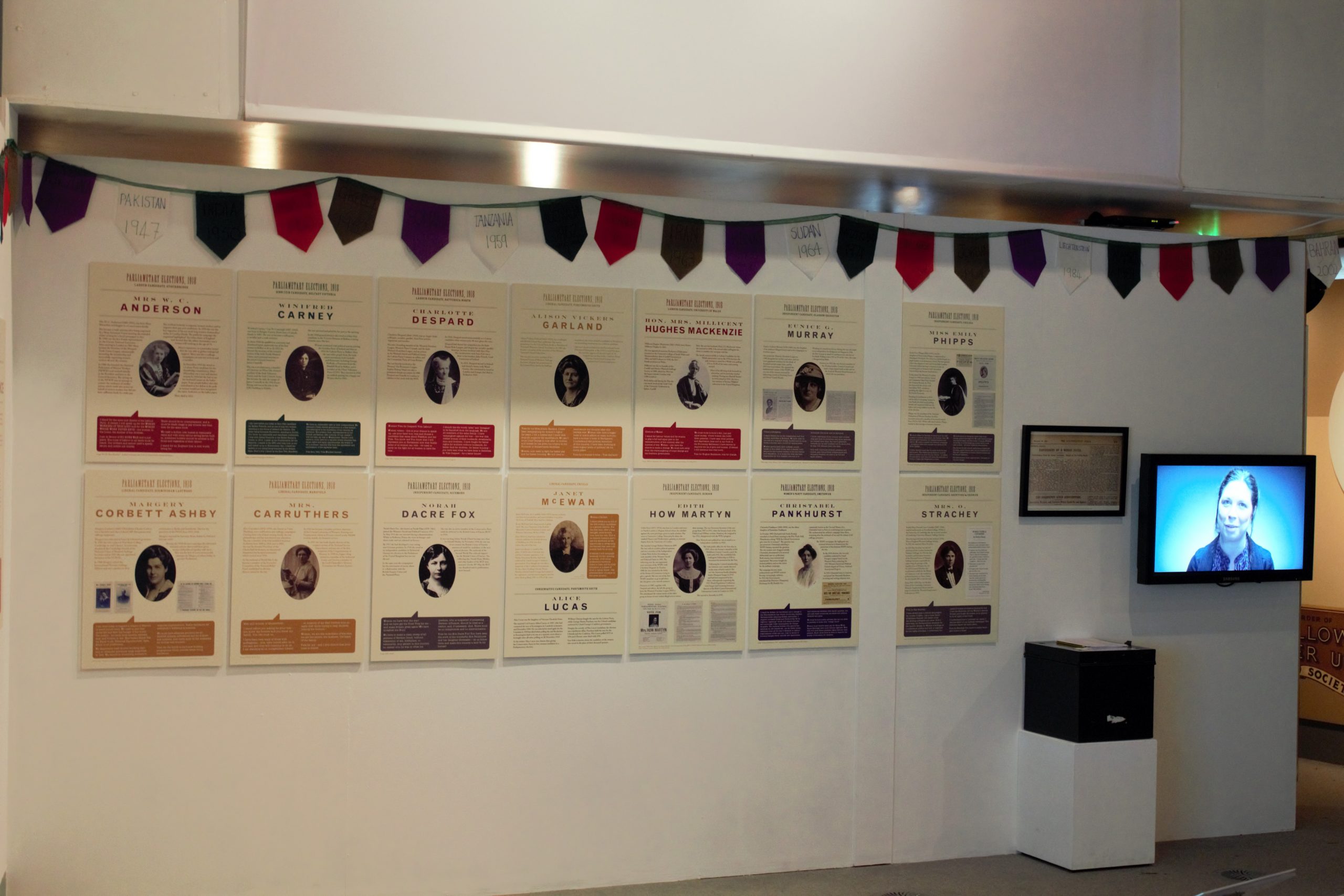 The women who said 'yes'! exhibition at People's History Museum, 2018. Photograph of a long exhibition wall with 14 large panels that have text and images on. At the right end of the wall there is a television monitor and a framed document. The top of the wall is decorated with colourful triangular bunting.