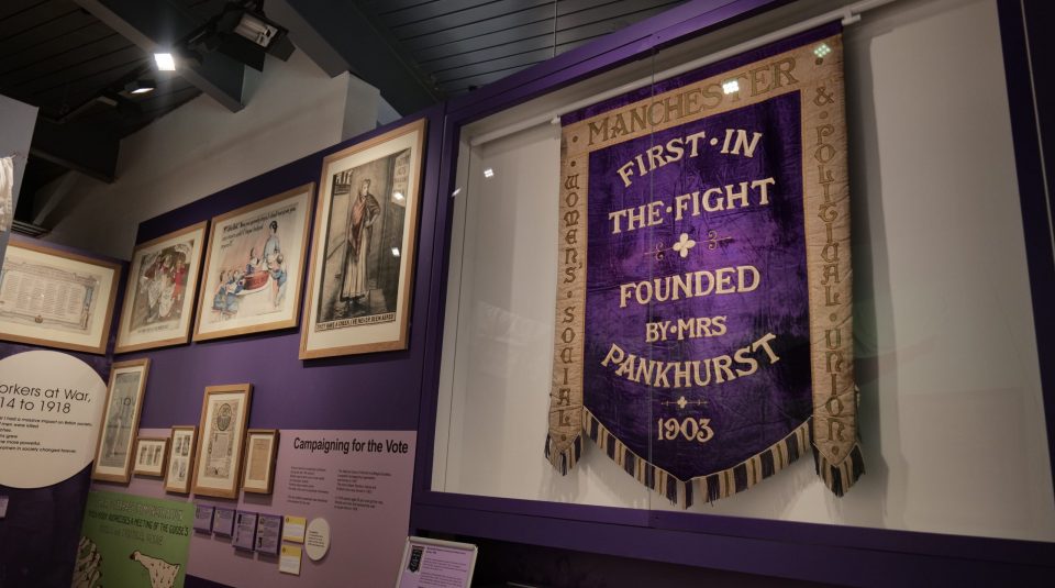 Image of Purple banner with the words ‘First in the fight. Founded by Mrs Pankhurst. 1903.’ There is a gold border with the word ‘Manchester’ at the top, ‘Women’s social’ on the left side and ‘& political union’ on the right. 