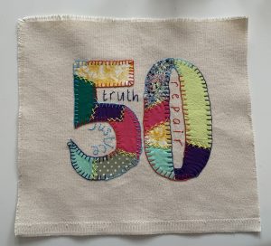 Colorful patchwork '50' stitched onto the center of the fabric square with the words 'truth', repair' and 'justice stitched around it.