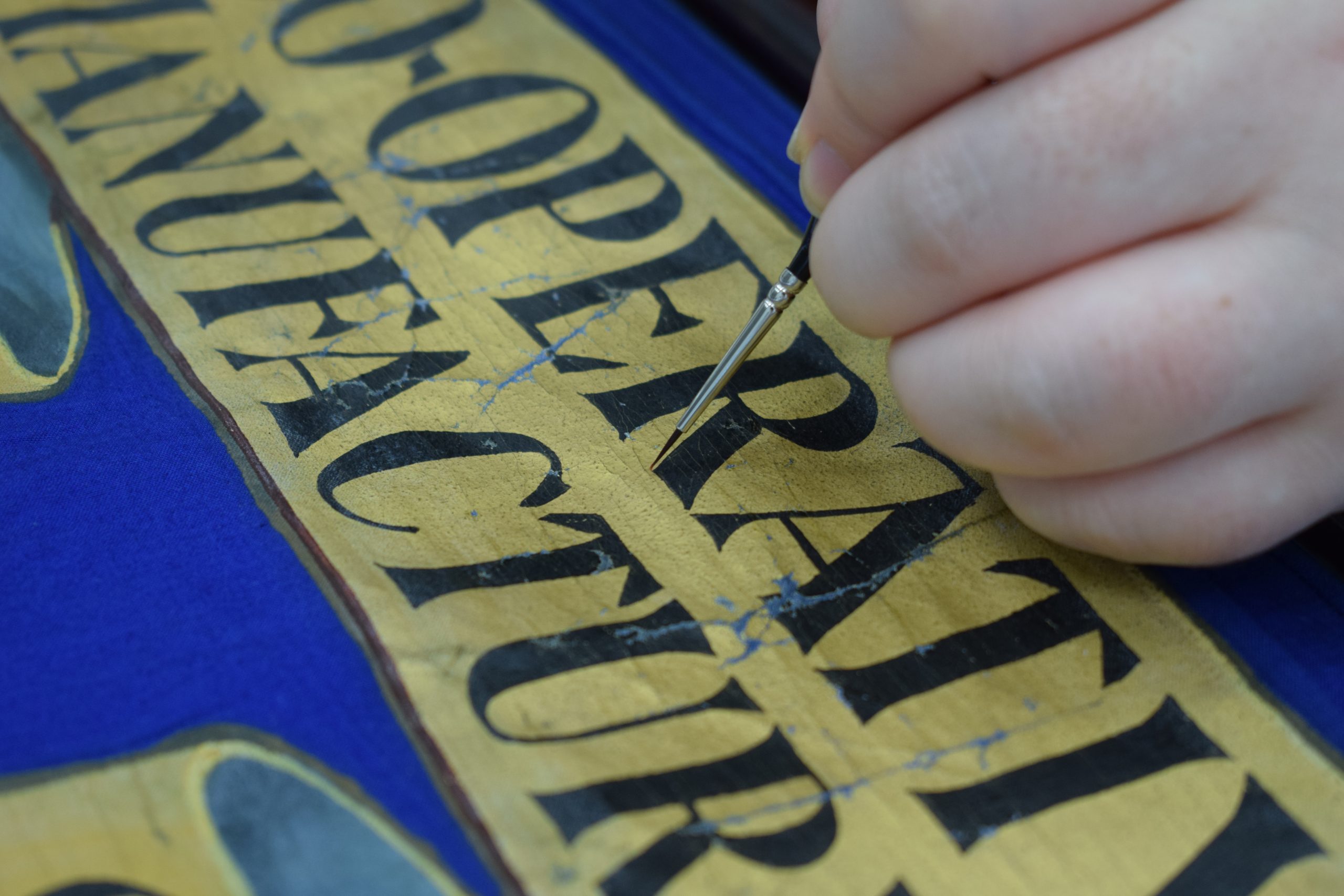 A person’s hand holding a very thin paintbrush over the painted surface of a banner.
