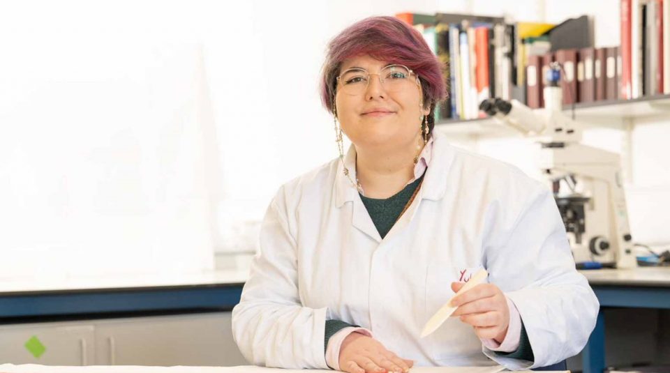 Image of A photo of the author in a lab coat. They have round glasses, pinky-purple hair, and are smiling.