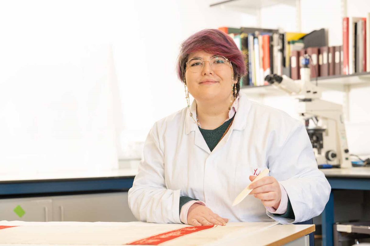 A photo of the author in a lab coat. They have round glasses, pinky-purple hair, and are smiling.