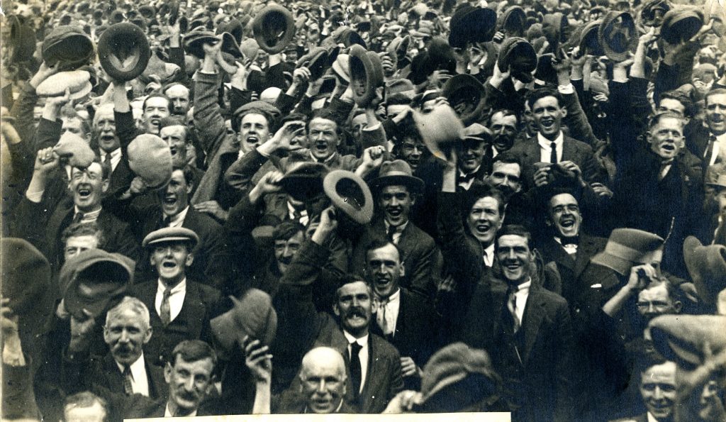 Photograph of a large group of people in suits cheering and raising their hats and caps