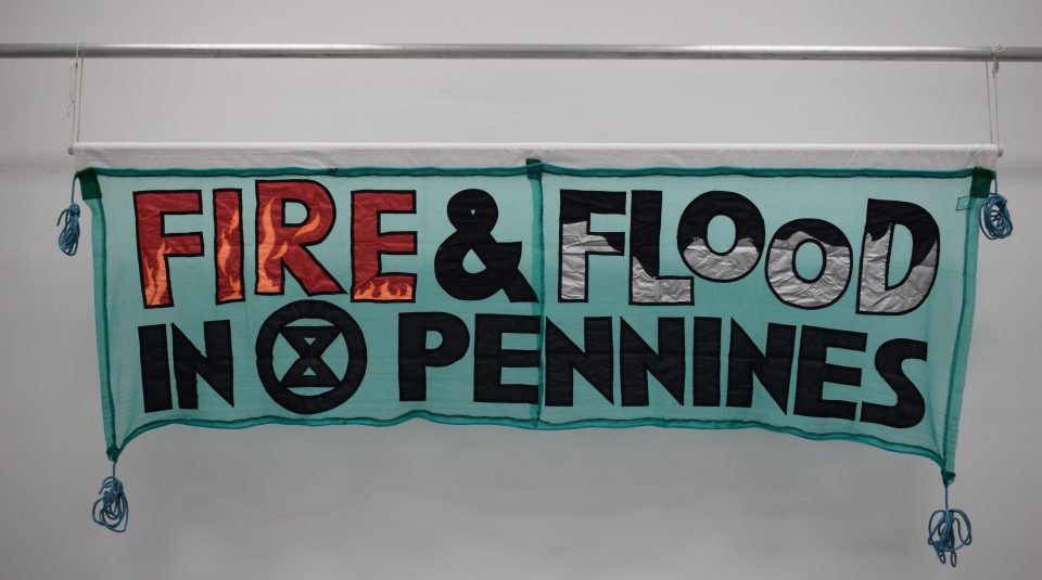 Image of blue landscape banner with the words ‘FIRE & FLOOD IN PENNINES’. The word ‘FIRE’ is in red, with light orange flames. The word ‘FLOOD’ is in black, with grey waves. The remaining words are black. Each corner of the banner has a tied knot of blue rope hanging.