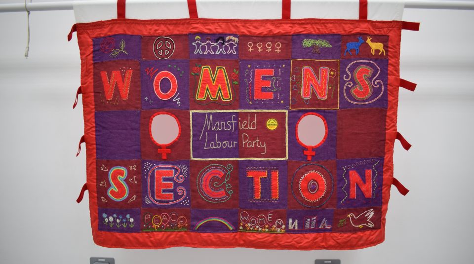 Image of Landscape banner made of burgundy and purple fabric squares in a patchwork pattern, framed by a red border. Decorated by needlework imagery including red roses, white doves, and different coloured shapes, with text 'Mansfield Labour Party Women’s Section'.