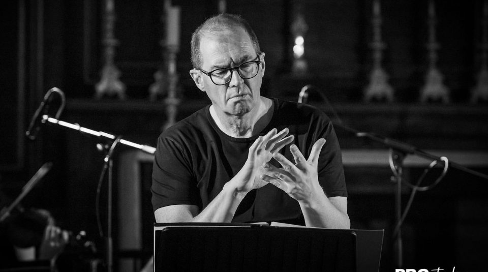 Image of black and white image of Paul Whittaker of Manchester's Hand Made Signing Choir conducting the choir using British Sign Language (BSL) interpretation. He is a white man wearing dark rimmed glasses and a black T-shirt, standing behind a lectern.