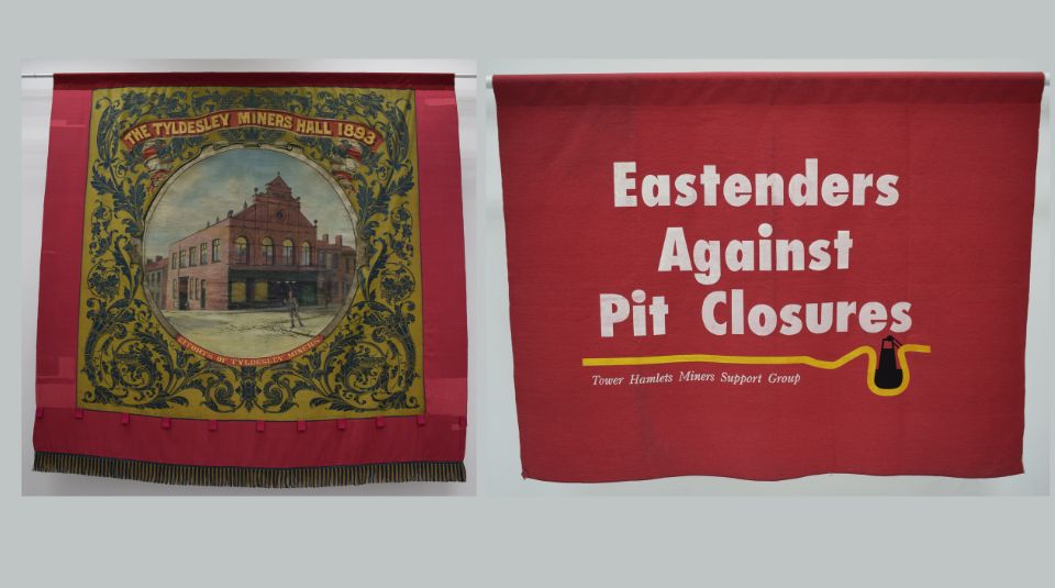 left to right: red bordered banner with central image of a brick building with text: The Tyldesley Miners’ Hall, 1893' and a red banner with text: 'Eastenders Against Pit Closures, Tower Hamlets Miners Support Group'.