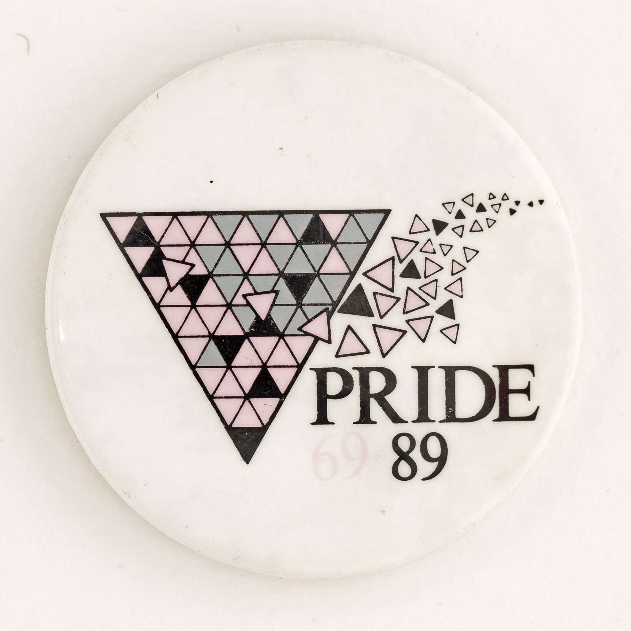 A white badge with a downwards facing triangle filled with lots of smaller pink, grey, and black triangles. Text reads 'Pride 69 89'.