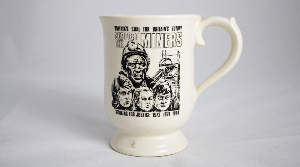 Image of A white mug with black line drawings of a miner and family with the text Support the Miners.