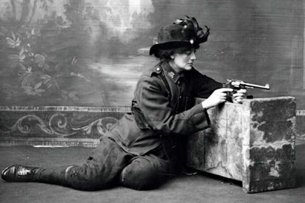 Constance Markievicz in uniform, kneeling against a studio prop holding a gun.