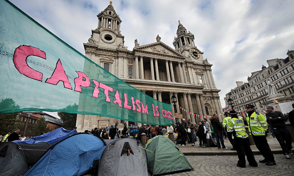 A large banner which says 'Capitalism Is Crisis' flying outside St Paul's Cathedral, London, above a cluster of tents on the left and police officers on the right.
