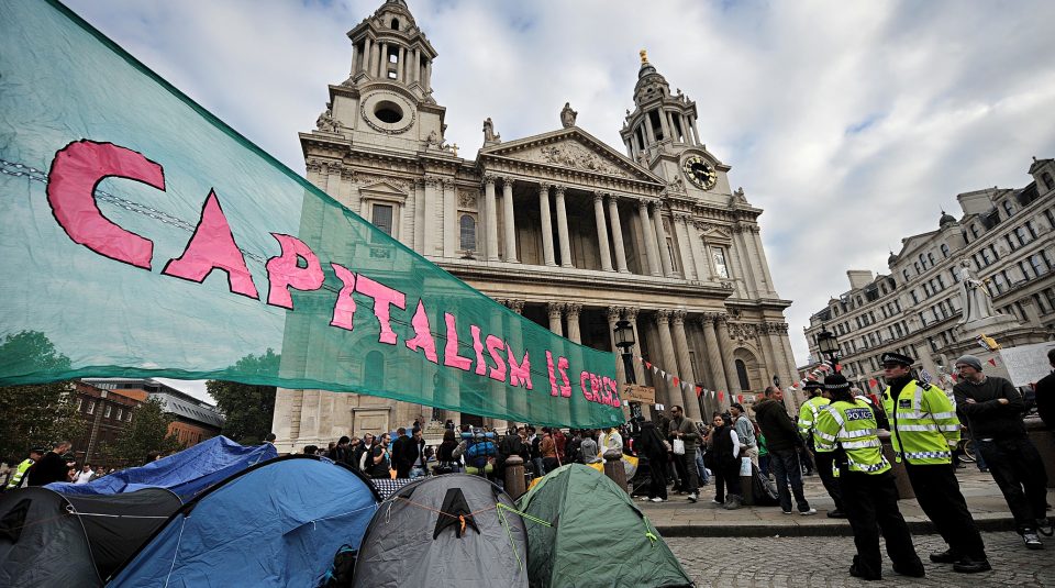 Image of A large banner which says 'Capitalism Is Crisis' flying outside St Paul's Cathedral, London, above a cluster of tents on the left and police officers on the right.
