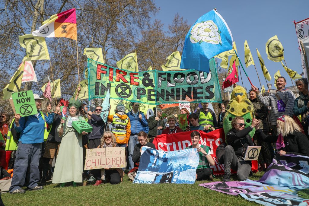Image of Lots of people gathered outside on a sunny day, holding up climate activism banners and flags. In the centre is a large blue 'netty' banner which says 'Fire & Flood In Pennines' with the Extinction Rebellion logo.