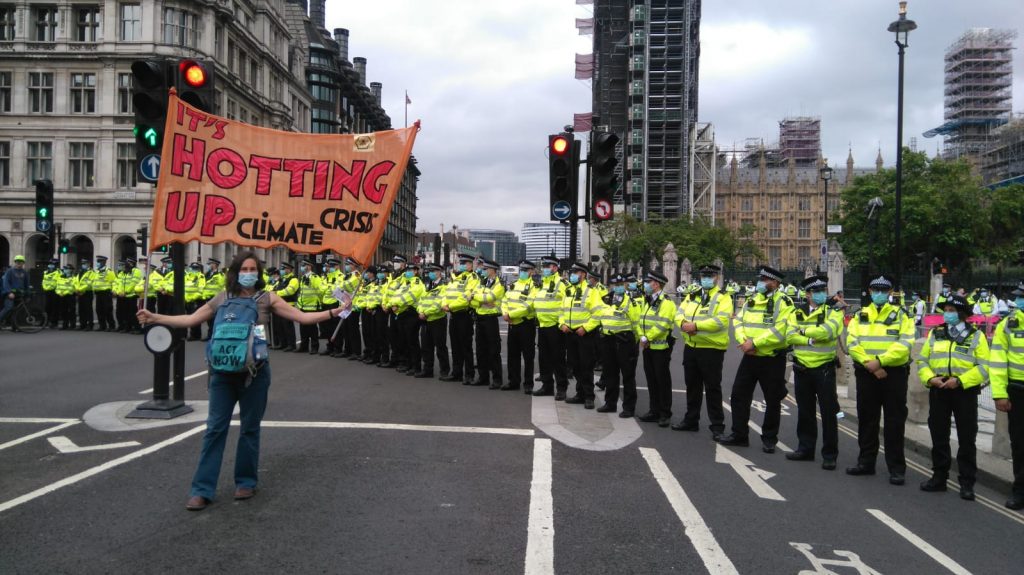A person in the middle of an empty road holding up an 'It's Hotting Up Climate Crisis' banner in front of a long line of police officers.