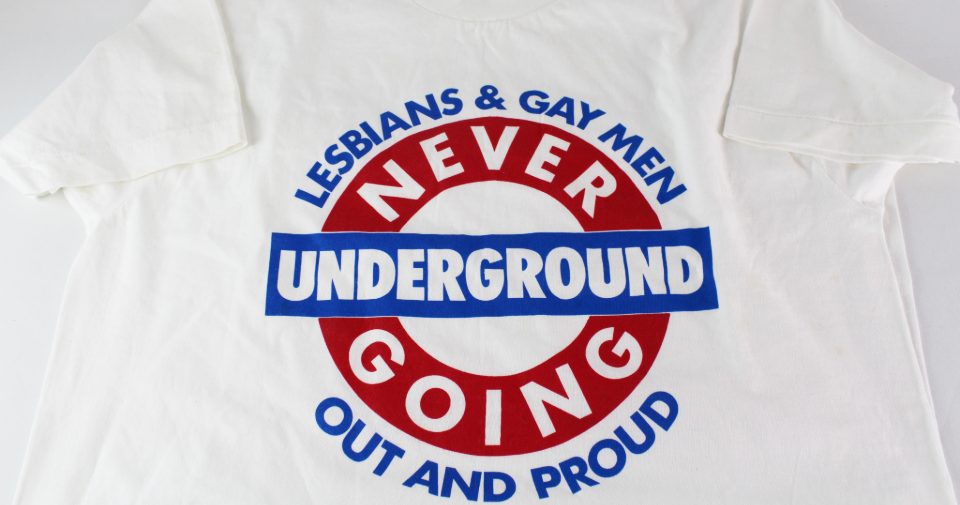 Image of white t-shirt with blue and red London Underground style logo with text 'Lesbians & Gay Men, Never Going Underground, Out And Proud'.