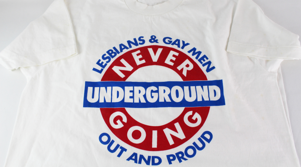 Image of white t-shirt with blue and red London Underground style logo with text 'Lesbians & Gay Men, Never Going Underground, Out And Proud'.