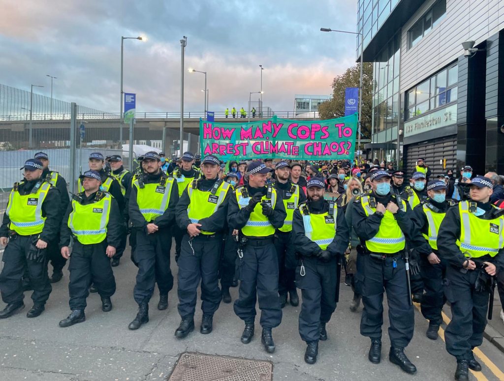 A line of police officers in front of a large group of protestors holding a 'How Many Cops To Arrest Climate Chaos?' banner.