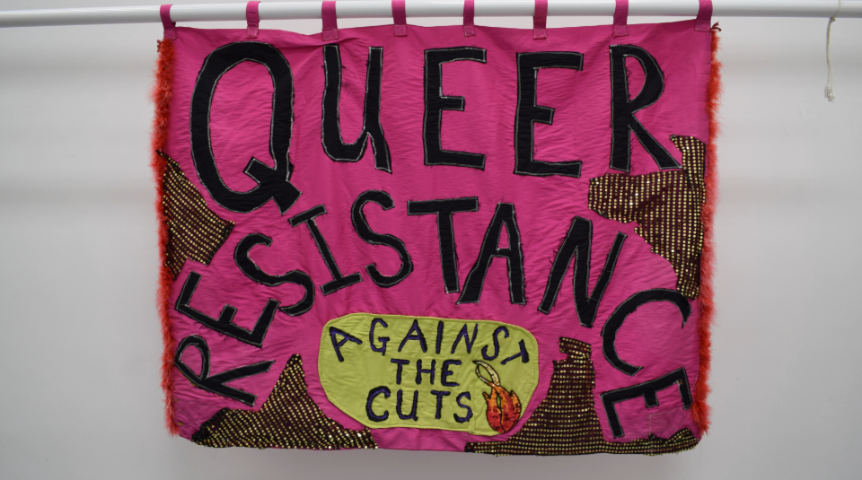 Image of a pink banner with glitter paint and red feather trim on both vertical sides, with black text reading: 'Queer Resistance Against The Cuts'.