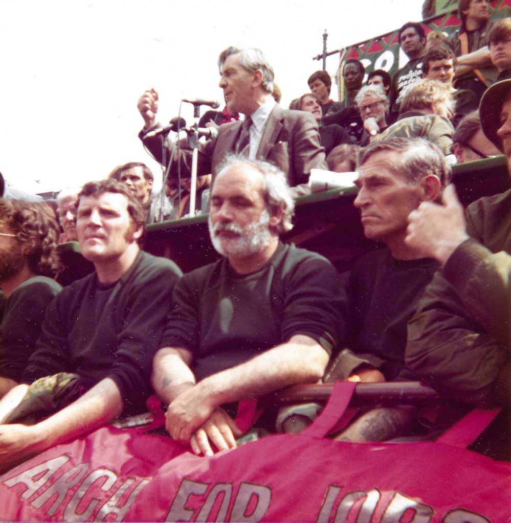 A group of people sitting with a banner in a stadium. Behind them is a person speaking into a microphone, with their fist raised.