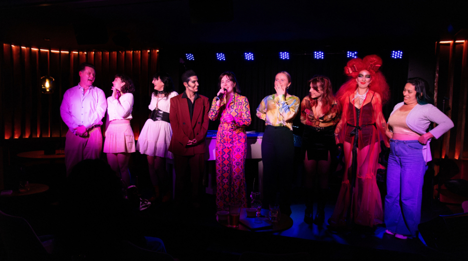 Image of Colour photogragh of nine performers from Queens of the Desert on stage. They look celebratory and are smiling.
