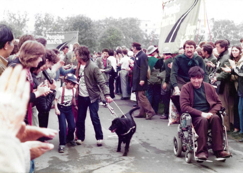 Colour photograph of a person with a dog and a person in wheelchair with a group of people clapping at a protest march. There are two banners with the text: 'People’s march for jobs'.
