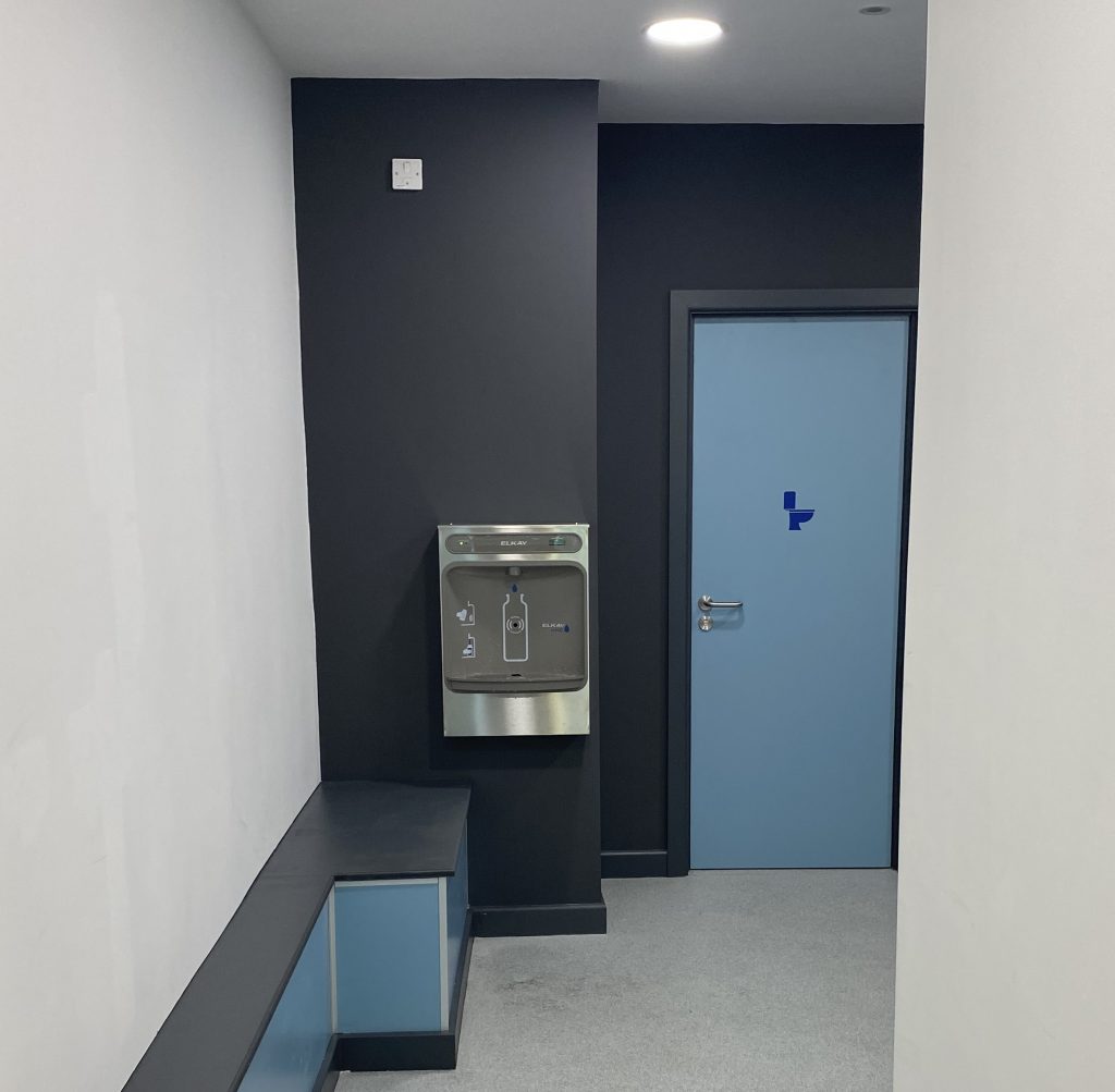 bottle filling station on a dark grey wall, next to a light blue doorway with a dark blue symbol for a toilet on.