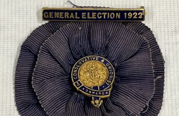 Image of Navy blue fabric rosette with a blue and gold badge in the centre with text reading: 'Conservative & Unionist, Onward' and a blue and gold pin at the top reading: 'General Election 1922'.