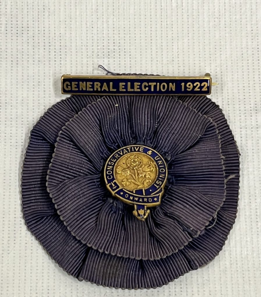 Navy blue fabric rosette with a blue and gold badge in the centre with text reading: 'Conservative & Unionist, Onward' and a blue and gold pin at the top reading: 'General Election 1922'.