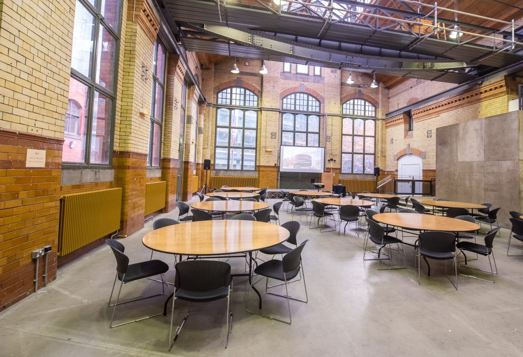 a large space with exposed brick walls and large windows, with several round tables with chairs around them.