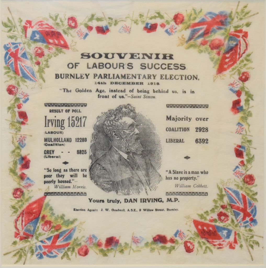 Napkin bordered with red roses and flags, with a central illustration of Dan Irving MP, and black text including: 'Souvenir of Labour's Success, Burnley Parliamentary Election, 14th December 1918.'