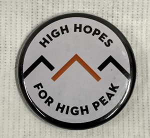 A circular badge with a black trim and white background, with three triangular 'peaks' in black, red, and black, with black text: 'High Hopes For High Peak'.