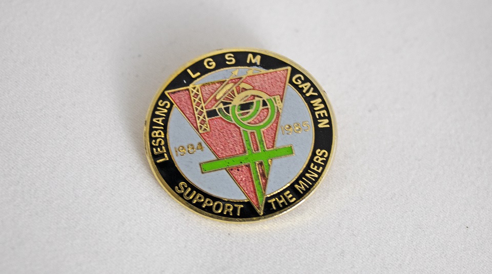 Image of A round badge with an upside down pink triangle superimposed with a gold pit wheel and a green female symbol, on a blue background. The edge of the badge is black with gold lettering: ‘LGSM. Lesbians. Gay Men. Support the Miners. 1984. 1985.’
