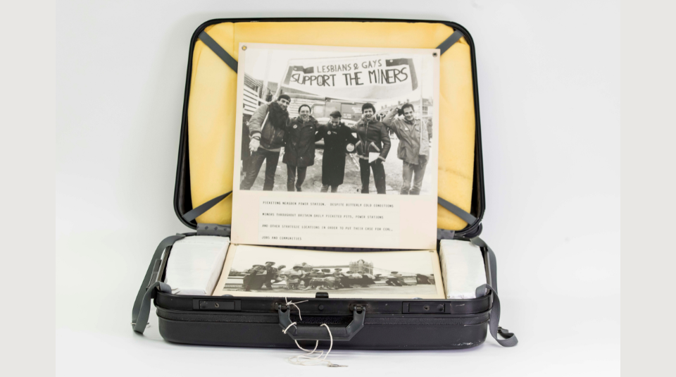 Image of An open black suitcase containing boards from a photographic exhibition, displaying a black and white photograph of five people standing under a banner which reads: ‘Lesbians & Gays Support The Miners’.