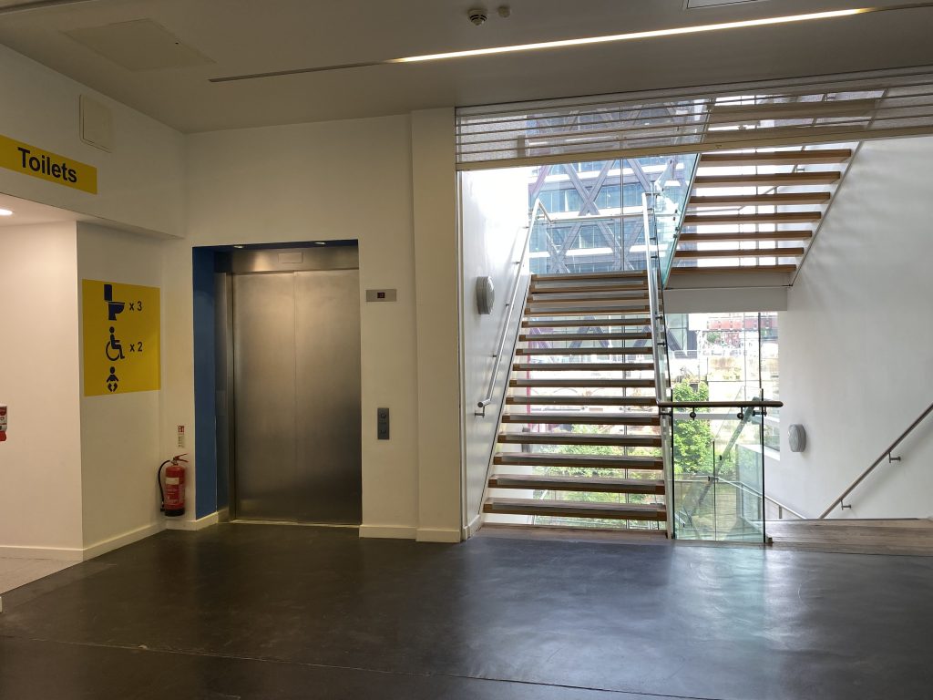 left to right: entrance to toilets, lift doors, a staircase leading up, and a staircase leading down.