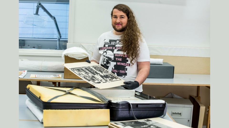 Image of PHM Collections Assistant Jaime Starr wearing a white t-shirt with an advertisement for the LGSM Pits and Perverts club night on it in black and pink, including a miner’s face in profile. Jaime is holding a black and white photo from the LGSM exhibition suitcase, also pictured.