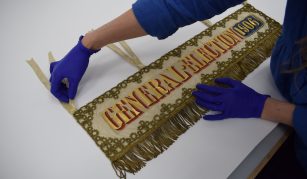 Image of Conservator's gloved hands handling a landscape banner with gold fringing, embroidery, and lettering reading: 'General Election 1906'.
