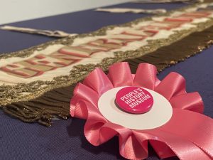 Pink rosette with a People's History Museum badge. Behind is a landscape banner with gold fringing, embroidery, and lettering reading: 'General Election 1906'.