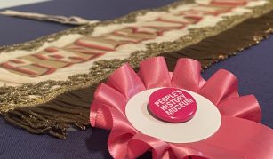 Image of Pink rosette with a People's History Museum badge. Behind is a landscape banner with gold fringing, embroidery, and lettering reading: 'General Election 1906'.