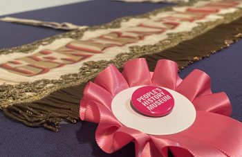 Image of Pink rosette with a People's History Museum badge. Behind is a landscape banner with gold fringing, embroidery, and lettering reading: 'General Election 1906'.