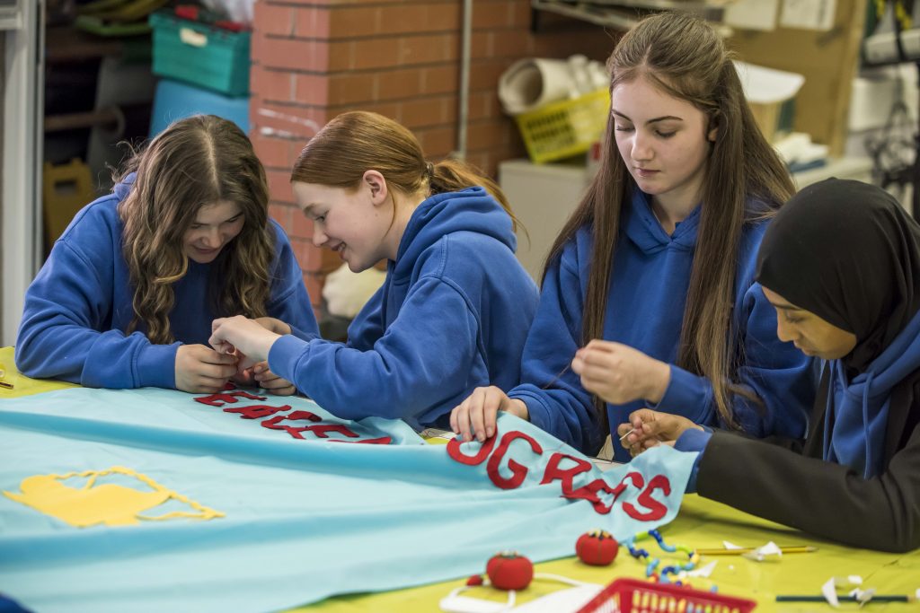 Three school students stitching red letters onto a blue banner