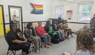 Image of The image shows seven seated people facing an audience. The wall behind them has a rainbow colour pride progress flag on it.