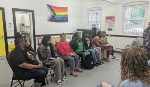 Image of The image shows seven seated people facing an audience. The wall behind them has a rainbow colour pride progress flag on it.